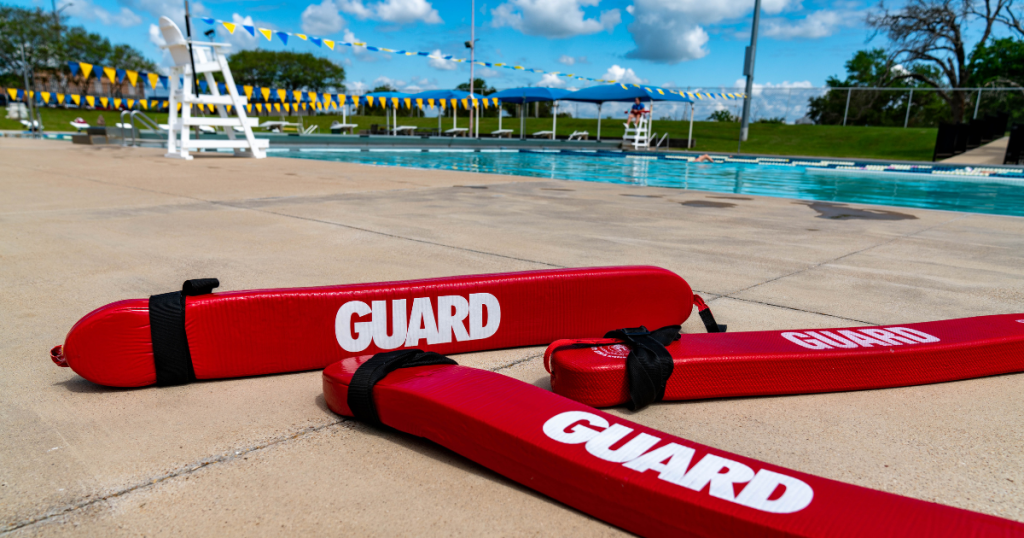 Three red "GUARD" labeled rescue tubes placed prominently in front of an outdoor swimming pool on a sunny day with clear skies and a lifeguard chair in the background.