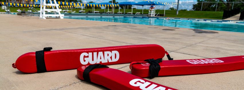 Three red "GUARD" labeled rescue tubes placed prominently in front of an outdoor swimming pool on a sunny day with clear skies and a lifeguard chair in the background.