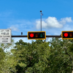 Traffic light intersection with signs indicating "LEFT TURN YIELD ON FLASHING YELLOW ARROW," and lane directions for straight and right turns only, set against a backdrop of trees and blue sky.