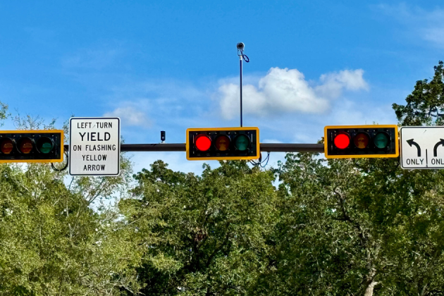 Traffic light intersection with signs indicating "LEFT TURN YIELD ON FLASHING YELLOW ARROW," and lane directions for straight and right turns only, set against a backdrop of trees and blue sky.