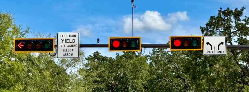 Traffic light intersection with signs indicating "LEFT TURN YIELD ON FLASHING YELLOW ARROW," and lane directions for straight and right turns only, set against a backdrop of trees and blue sky.